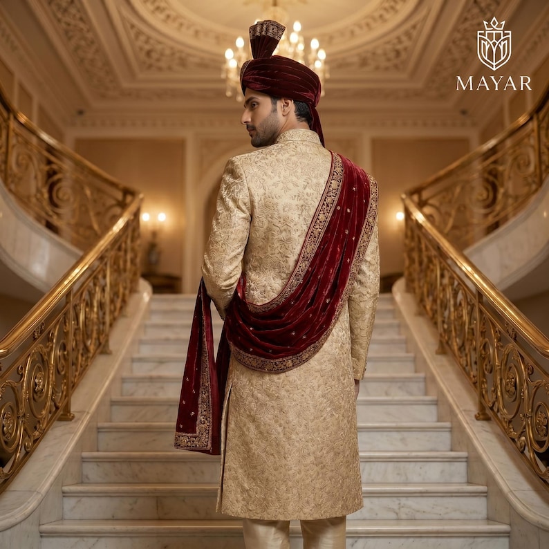 May include: A gold sherwani featuring intricate tonal embroidery, paired with a deep red velvet turban and a matching red velvet shawl draped over the shoulder. The garment includes a high collar and long sleeves, showcasing a detailed pattern.