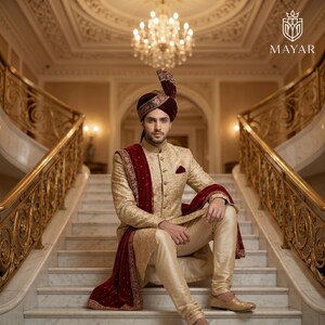 May include: A gold sherwani with intricate embroidery, paired with matching trousers and a deep red velvet stole. The ensemble includes a matching maroon turban with gold detailing. The Mayar logo is visible in the upper right corner.