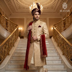 May include: A gold sherwani with intricate embroidery, a matching turban, and a deep red velvet stole. The garment features a high collar, a button-down front, and a pocket square. The Mayar logo is visible in the top right corner of the image.