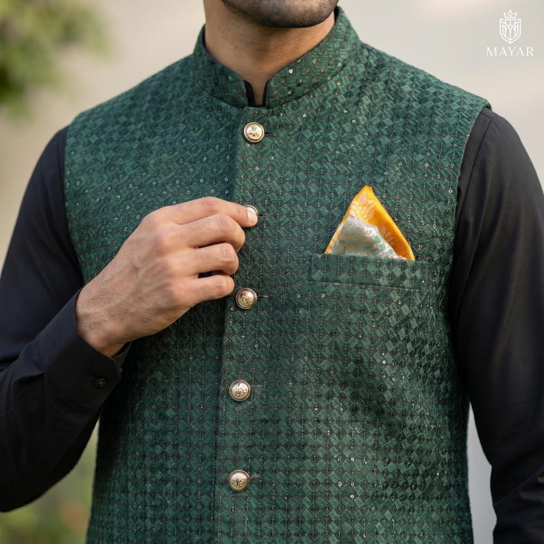 May include: A dark green vest with a geometric pattern and gold buttons. The vest has a mandarin collar and a pocket with a yellow and green patterned pocket square. The vest is worn over a black long-sleeve shirt. The Mayar logo is in the top right corner.