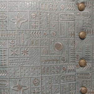 K&ouml;nnte beinhalten: Nahaufnahme einer hellblauen Jacke mit aufwendigen Stickmustern. Die Jacke weist eine Reihe geometrischer und floraler Designs in wei&szlig;em Faden auf, mit goldfarbenen Kn&ouml;pfen. Die Gesamt&auml;sthetik ist detailliert und elegant.