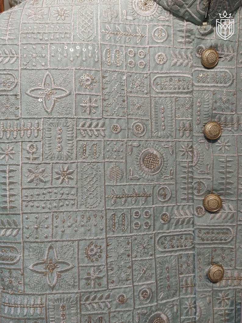 K&ouml;nnte beinhalten: Nahaufnahme einer hellblauen Jacke mit aufwendigen Stickmustern. Die Jacke weist eine Reihe geometrischer und floraler Designs in wei&szlig;em Faden auf, mit goldfarbenen Kn&ouml;pfen. Die Gesamt&auml;sthetik ist detailliert und elegant.