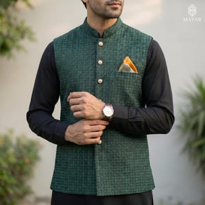 May include: A green textured vest with gold buttons and a pocket square. The vest is worn over a black long-sleeve shirt. A wristwatch with a brown leather band is visible. The vest has a mandarin collar.
