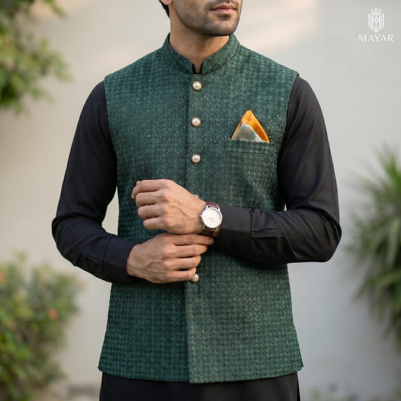 May include: A green textured vest with gold buttons and a pocket square. The vest is worn over a black long-sleeve shirt. A wristwatch with a brown leather band is visible. The vest has a mandarin collar.