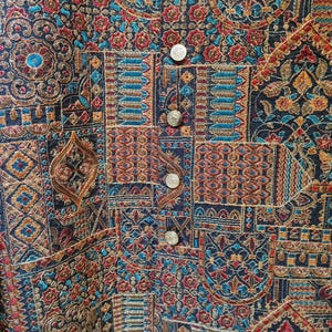 May include: Close-up of a detailed, embroidered vest with a patchwork design. The vest features a rich tapestry of colors including navy blue, red, teal, and gold. Silver buttons run down the front. The intricate patterns suggest a cultural or vintage aesthetic.