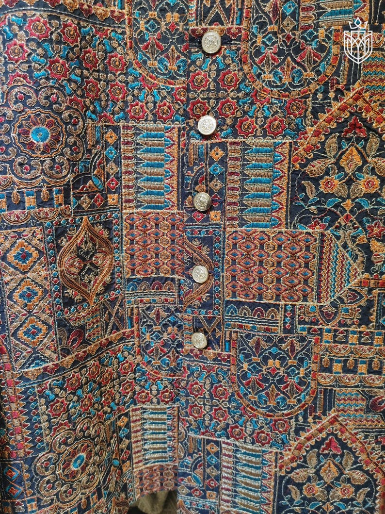 May include: Close-up of a detailed, embroidered vest with a patchwork design. The vest features a rich tapestry of colors including navy blue, red, teal, and gold. Silver buttons run down the front. The intricate patterns suggest a cultural or vintage aesthetic.