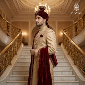 May include: A beige sherwani featuring intricate gold embroidery, paired with a deep maroon velvet turban and a matching maroon velvet shawl draped over the shoulder. The garment includes a pocket square and buttons, with the Mayar logo visible.