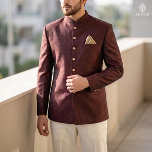 May include: A dark maroon bandhgala jacket featuring a subtle tonal pattern, a mandarin collar, and gold-toned buttons down the front. The jacket includes a chest pocket with a beige pocket square and long sleeves with gold-toned button cuffs.
