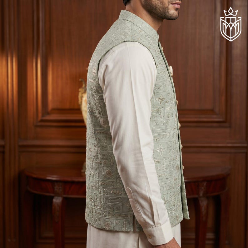 May include: A light green vest with an intricate embroidered pattern, worn over a white long-sleeved shirt. The vest has a mandarin collar and gold-colored buttons. The outfit is part of a formal wear collection.