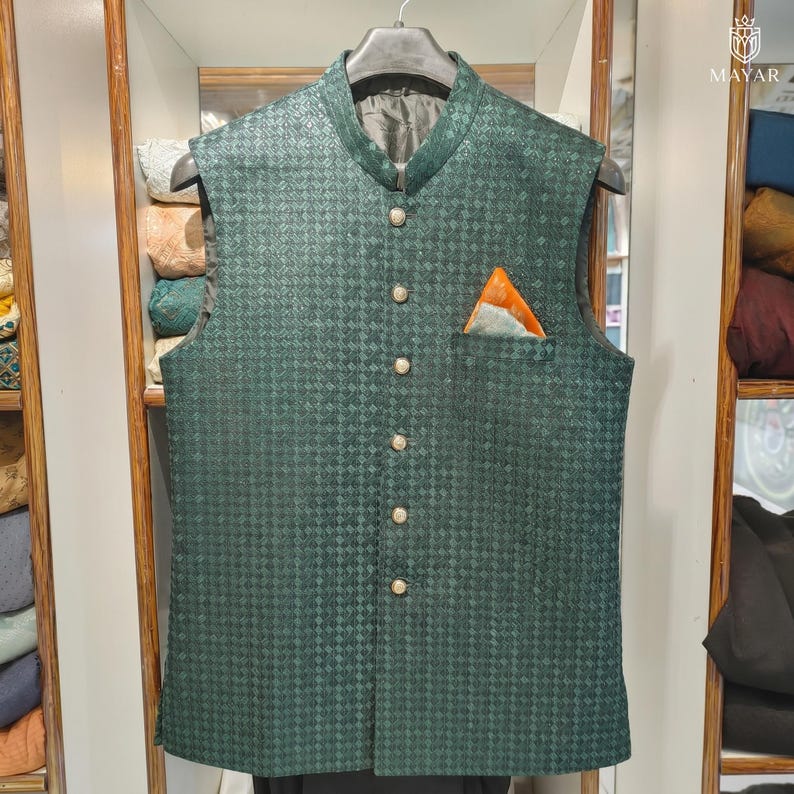 May include: A dark green vest with a textured, checkered pattern. The vest has a mandarin collar, gold-colored buttons, and a pocket square with orange and blue accents. The vest is sleeveless and hangs on a black hanger.