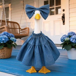 May include: A white goose figurine with a yellow beak and orange feet, wearing a blue denim dress with a gathered neckline and a matching oversized blue denim bow headband. The figure stands upright, showcasing the textured fabric of the outfit.