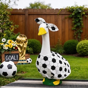 May include: A whimsical goose statue with a white body, yellow beak, and soccer ball-themed accents. The goose wears a soccer ball patterned outfit and headpiece. A soccer ball and a golden trophy are in the background.