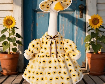 Pode incluir: Uma estatueta de ganso branco vestindo um vestido amarelo e um chapéu de sol a condizer, ambos com um padrão de girassóis. O vestido tem mangas bufantes, uma fita castanha no colarinho e um acabamento em renda branca na bainha.