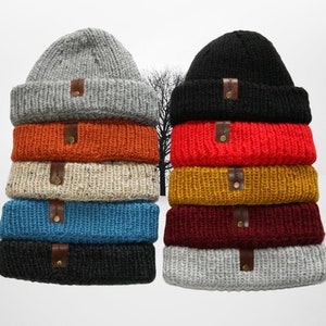 May include: A stack of ten knitted beanies in various colours, including grey, black, orange, beige, blue, burgundy, and yellow. Each beanie has a leather patch with the word "Handmade" on it.