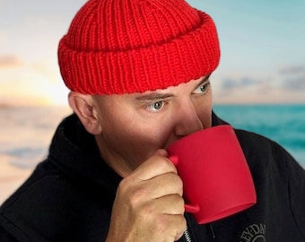 Könnte beinhalten: Eine Person trägt eine rote Strickmütze und eine schwarze Jacke und trinkt aus einer roten Tasse.