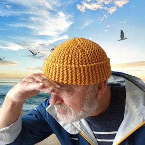 May include: A man wearing a yellow knitted beanie and a blue jacket with a white and blue striped shirt underneath. He is standing on a beach with the ocean in the background.