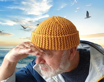 Könnte beinhalten: Ein Mann trägt eine gelbe Strickmütze und eine blaue Jacke mit einem weiß-blau gestreiften Hemd darunter. Er steht an einem Strand mit dem Meer im Hintergrund.