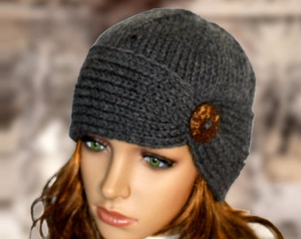 May include: A grey knitted cloche hat with a brown button accent. The hat has a slouchy fit and a wide brim.