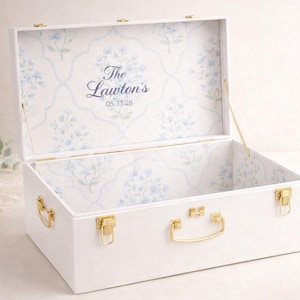 May include: A white rectangular storage chest with gold-toned metal latches and a handle. The interior is lined with a light blue floral pattern on a white background, featuring the text The Lawton's and the date 05.31.26 printed in the center.