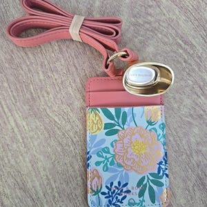 May include: A pink floral print lanyard with a matching cardholder. The cardholder features a light blue background with pink and gold floral designs. The lanyard is made of a soft, leather-like material and includes a gold-colored metal clasp.