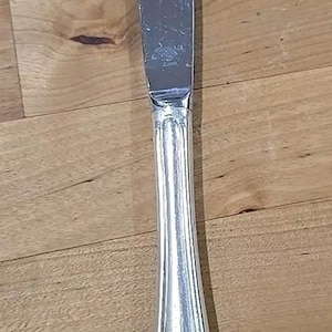 May include: A silver butter knife with a curved blade and a decorative handle. The handle has a fluted design and a floral motif at the base. The knife is made of a shiny, reflective metal.
