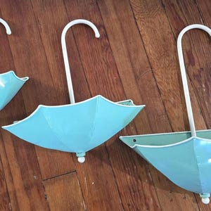 May include: Three light blue metal umbrella-shaped wall decorations with white trim and handles. The umbrellas are different sizes and are arranged on a wooden surface. The handles are curved.