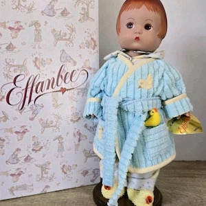 May include: A vintage Effanbee doll wearing a light blue corduroy robe with yellow trim and a yellow duck in the pocket. The doll has red hair and is standing on a wooden base. The box behind the doll has the Effanbee logo.