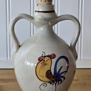 May include: Cream-colored ceramic jug with two handles and a cork stopper. The jug features a hand-painted rooster design in yellow, blue, and brown. The word "BOLLA" is printed on the cork. The jug sits on a wooden surface.