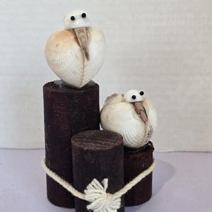 May include: A handcrafted decorative sculpture featuring two bird figures made from seashells. The birds are perched on dark brown wooden cylinders tied together with a white rope. The seashells are white and beige.