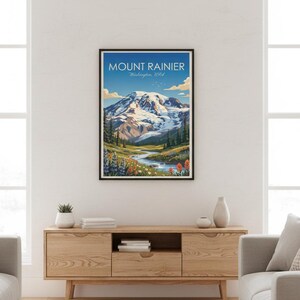 May include: Framed art print of Mount Rainier in Washington, USA. The artwork depicts a snow-capped mountain, a river, and colorful wildflowers under a blue sky. The print is in a black frame and is displayed on a wooden cabinet.