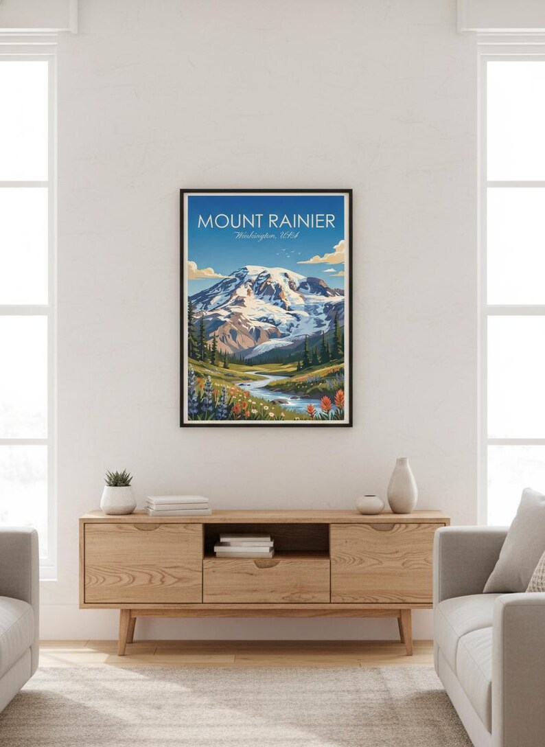 May include: Framed art print of Mount Rainier in Washington, USA. The artwork depicts a snow-capped mountain, a river, and colorful wildflowers under a blue sky. The print is in a black frame and is displayed on a wooden cabinet.