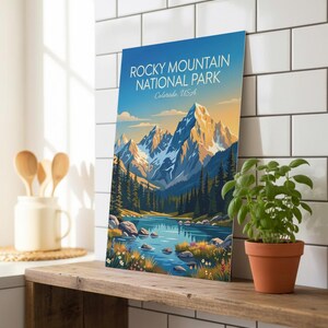May include: A framed print featuring the Rocky Mountain National Park in Colorado, USA. The image shows a mountain range with snow-covered peaks, a lake, and evergreen trees. The sky transitions from blue to yellow.