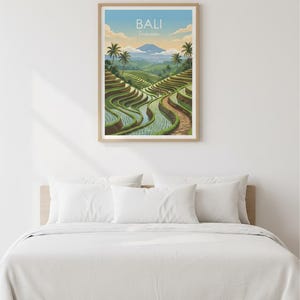 May include: A framed print featuring a scenic view of Bali, Indonesia. The artwork depicts lush green rice terraces, winding pathways, and palm trees under a blue sky. The print is displayed above a bed with white bedding.