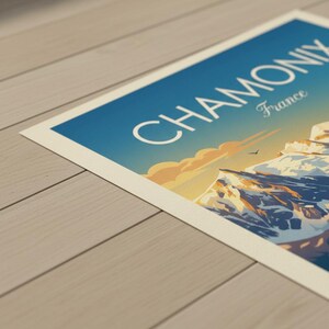 May include: A travel poster with the words "CHAMONIX France" set against a blue sky. The poster shows a mountain range with snowy peaks, a bird, and a warm sunset. The poster is displayed on a light-coloured wooden surface.