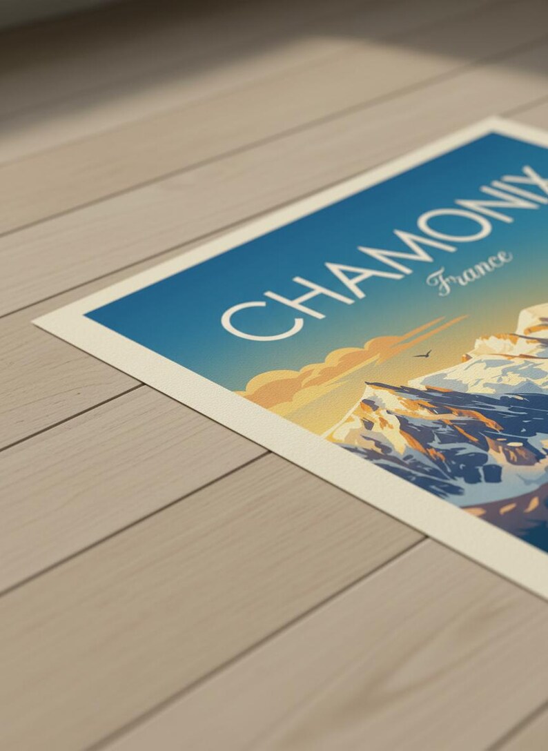May include: A travel poster with the words "CHAMONIX France" set against a blue sky. The poster shows a mountain range with snowy peaks, a bird, and a warm sunset. The poster is displayed on a light-coloured wooden surface.