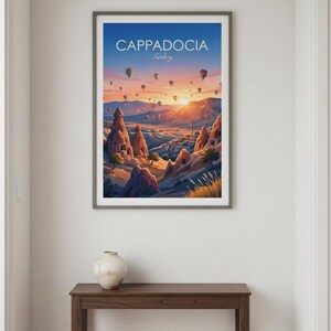 May include: Framed art print of Cappadocia, Turkey, featuring hot air balloons at sunrise. The artwork displays a vibrant landscape with unique rock formations, a town, and a colorful sky. The print is in a gray frame.