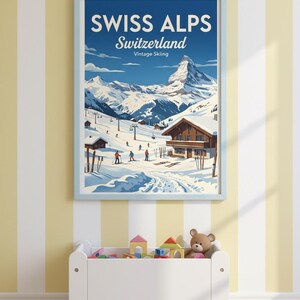 May include: A framed vintage poster of the Swiss Alps, featuring a snowy mountain landscape with skiers and chalets. The poster has a light blue frame and the text "SWISS ALPS Switzerland Vintage Skiing". Below, a white toy chest is filled with toys.