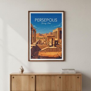 May include: A framed art print featuring a stylized illustration of Persepolis, Shiraz, Iran. The artwork depicts ancient ruins under a blue sky. The print is in a brown frame and is displayed on a wooden cabinet.