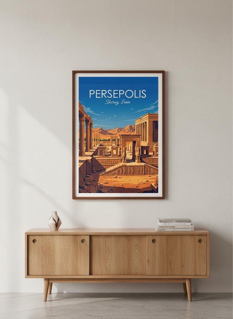 May include: A framed art print featuring a stylized illustration of Persepolis, Shiraz, Iran. The artwork depicts ancient ruins under a blue sky. The print is in a brown frame and is displayed on a wooden cabinet.
