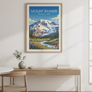 May include: A framed print of Mount Rainier in Washington, USA. The artwork depicts the snow-capped mountain under a blue sky, with a river flowing through a valley of wildflowers and evergreen trees. The print is displayed on a wooden console table.