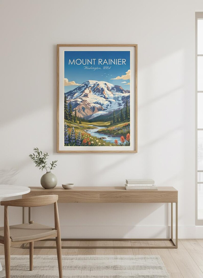 May include: A framed print of Mount Rainier in Washington, USA. The artwork depicts the snow-capped mountain under a blue sky, with a river flowing through a valley of wildflowers and evergreen trees. The print is displayed on a wooden console table.