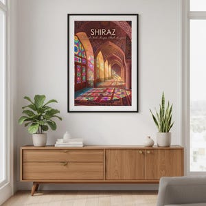 May include: A framed print depicting an arched corridor with stained glass windows and a vibrant carpet. The word "SHIRAZ" is at the top. The print is displayed on a wooden cabinet with plants.