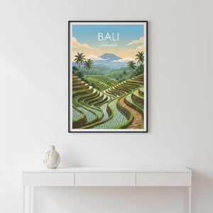 May include: A framed wall art print featuring a landscape illustration of Bali, Indonesia. The artwork depicts green terraced rice paddies, palm trees, and a distant mountain under a blue sky with clouds. The text BALI Indonesia is centered.