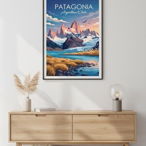 Op de afbeelding: Een ingelijste kunstafdruk met een landschap van Patagoni&euml;, Argentini&euml;/Chili. Het kunstwerk toont besneeuwde bergen, een blauw meer en goudkleurig gras onder een blauwe lucht. De tekst "PATAGONIA" is zichtbaar. De print hangt aan een houten kast.
