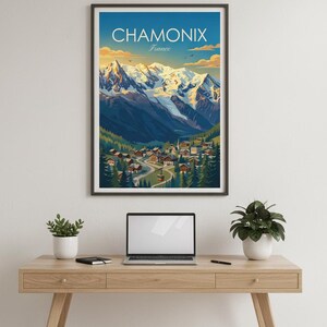 May include: A framed print of Chamonix, France, with the text "CHAMONIX France". The artwork shows a village in a valley with snow-covered mountains. The print is displayed on a wooden desk, with a laptop and plants. The print is in a black frame.