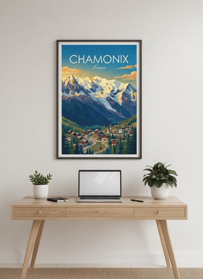 May include: A framed print of Chamonix, France, with the text "CHAMONIX France". The artwork shows a village in a valley with snow-covered mountains. The print is displayed on a wooden desk, with a laptop and plants. The print is in a black frame.
