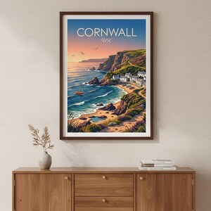 May include: A framed wall art print featuring a coastal landscape of Cornwall, UK. The illustration depicts a sunset over the ocean, a small sailboat, rocky cliffs, a sandy beach, and a cluster of white houses nestled along the green hillside.