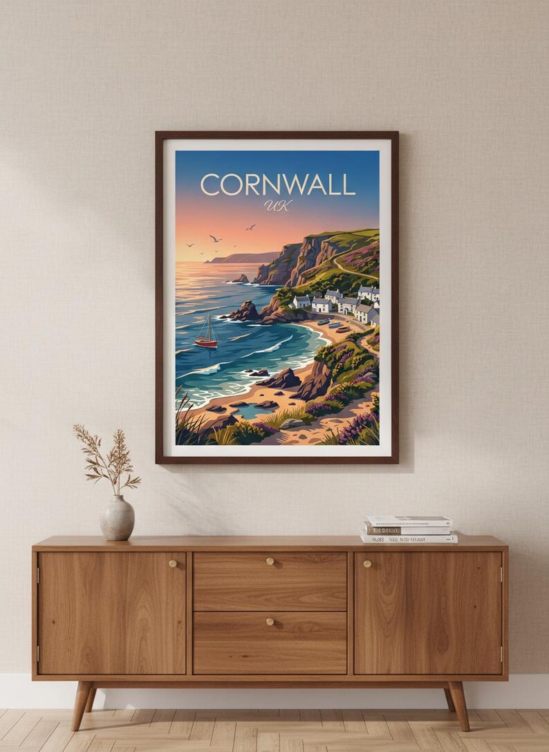 May include: A framed wall art print featuring a coastal landscape of Cornwall, UK. The illustration depicts a sunset over the ocean, a small sailboat, rocky cliffs, a sandy beach, and a cluster of white houses nestled along the green hillside.