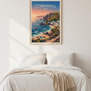 May include: A framed print featuring a coastal scene of Cornwall, UK. The artwork depicts a bay with a small village, cliffs, and a sailboat under a sunset sky. The print is displayed above a bed with white linens and a beige throw.