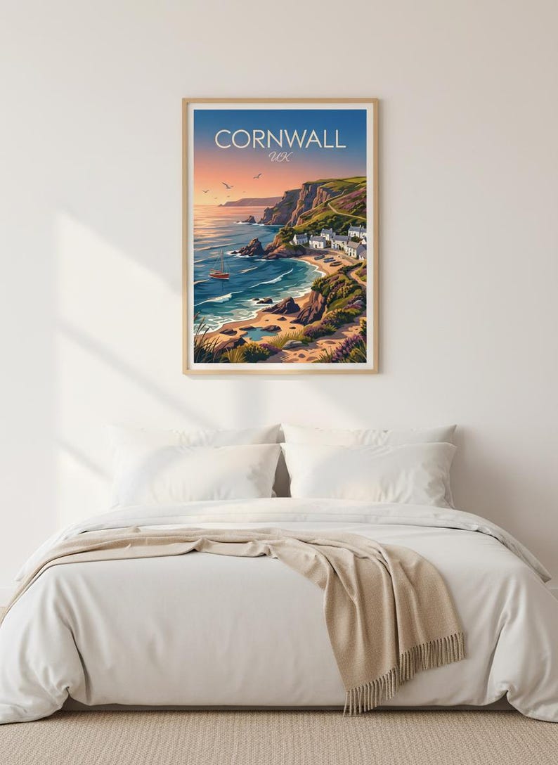 May include: A framed print featuring a coastal scene of Cornwall, UK. The artwork depicts a bay with a small village, cliffs, and a sailboat under a sunset sky. The print is displayed above a bed with white linens and a beige throw.